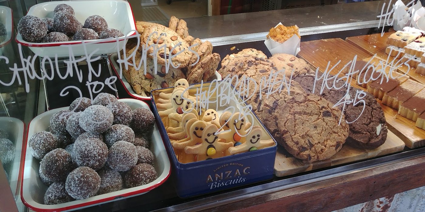 A glass cabinet shelf with chocolate rum balls, ANZAC biscuits in a tin, cookies and slices, with chalk price labels