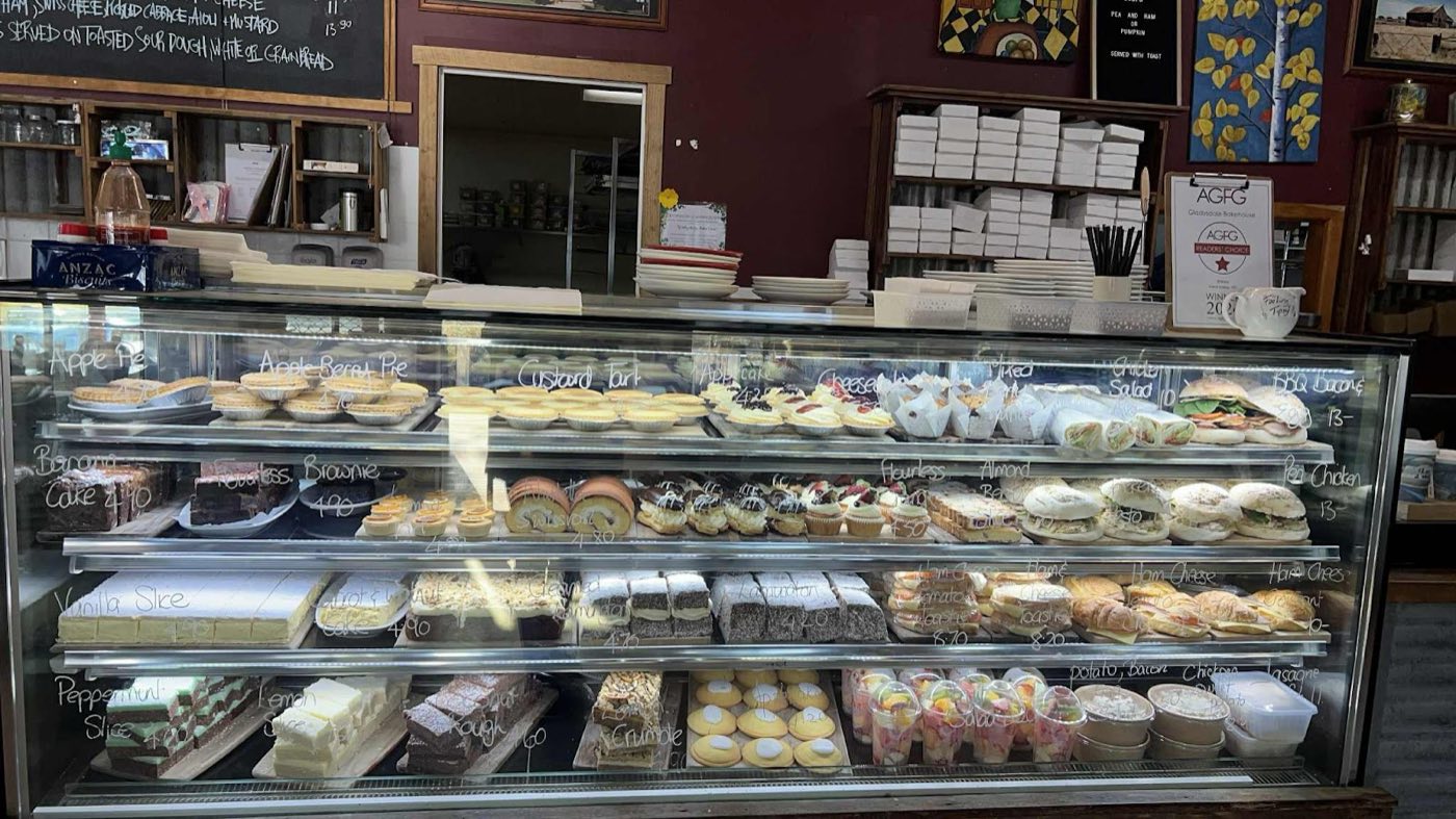 The long glass display cabinet full of pies, rolls, slices and cakes, with pendant lights and a corrugated-iron counter front