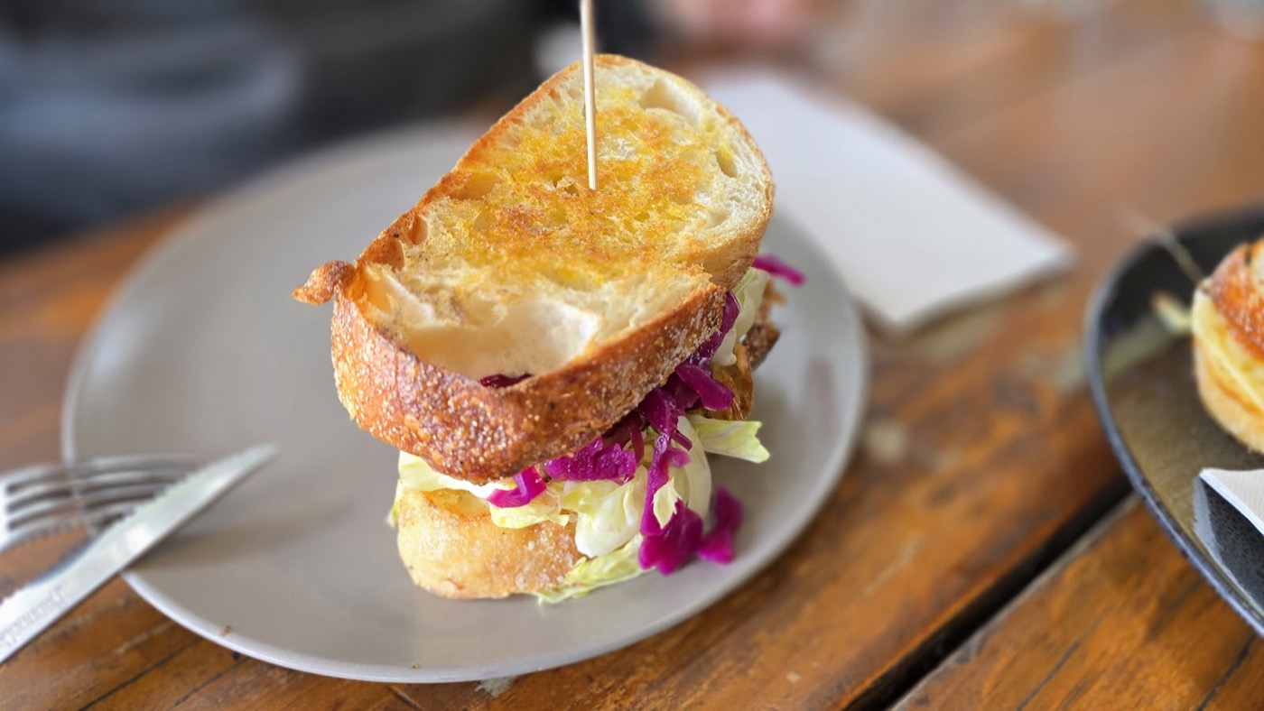 A toasted sandwich on golden crusty bread with purple cabbage and filling, held together with a skewer, on a grey plate