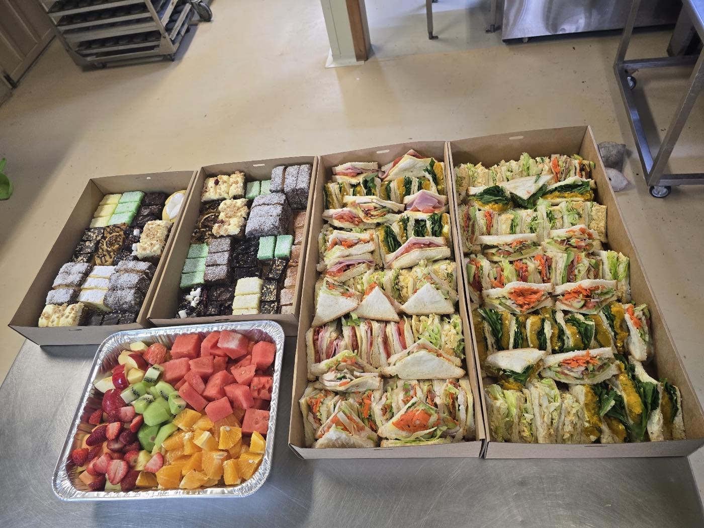 Trays of sandwiches, small slices and a tray of cut fruit laid out on a bench